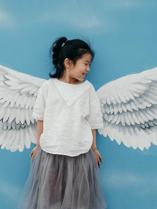 Ein kleines Mädchen steht lächelnd vor einer bemalten Wand: einem blauen Himmel, davor weisse Engelsflügel. Die Flügel sehen aus als wenn sie dem Mädchen angewachsen sind.
