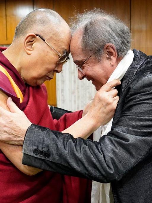 Der Dalai Lama, im roten Gewand, und der Theologe und Publizist Michael von Brück, im grauen Jackett, stehen einander zugewandt, Stirn an Stirn, und deuten eine Umarmung an.