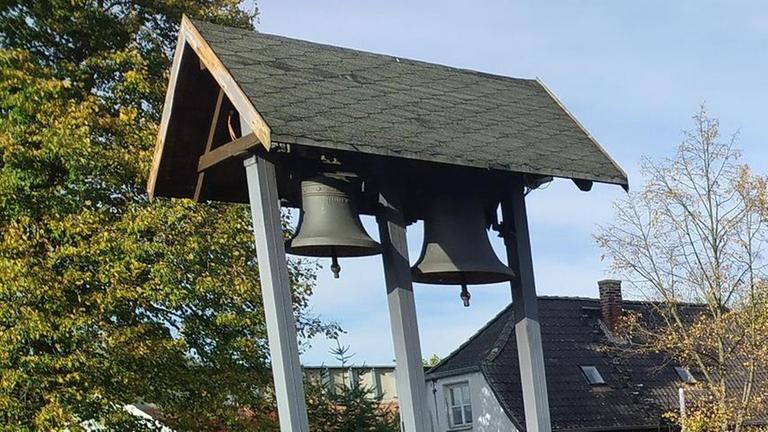 Zwei Glocken hängen in einem regengeschützten Holzgestell. Im Hintergrund sind Häuser der Gemeinde Jerche zu sehen.
