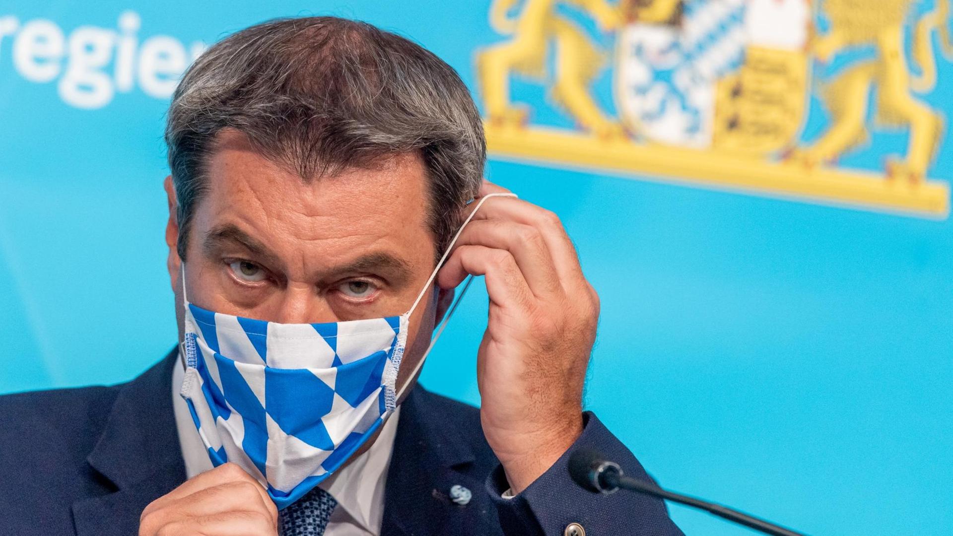 Söder vor einer blauen Wand mit dem bayerischen Wappen. Man sieht nur seinen Kopf. Der Maskenstoff zeigt die blau-weißen Rauten. Söder vor einer blauen Wand mit dem bayerischen Wappen. Man sieht nur seinen Kopf. Der Maskenstoff zeigt die blau-weißen Rauten.