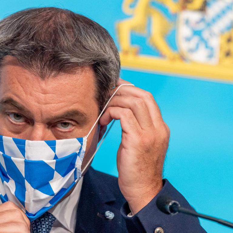 Söder vor einer blauen Wand mit dem bayerischen Wappen. Man sieht nur seinen Kopf. Der Maskenstoff zeigt die blau-weißen Rauten.  
