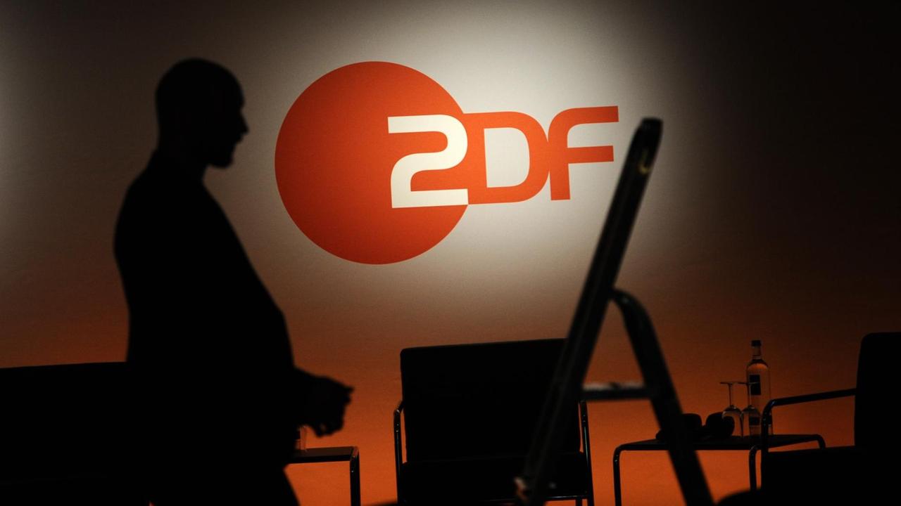 Ein Techniker steht neben einer Leiter vor einem ZDF-Logo. Auf einem Tisch sieht man Minerlwasser und Gläser.
