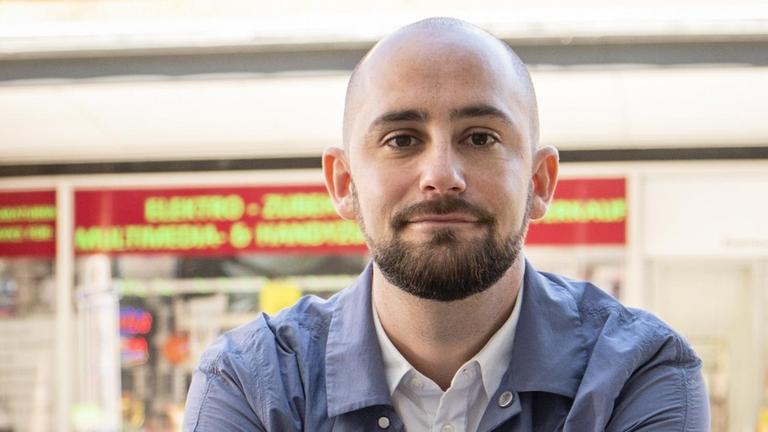 Philipp Staab, im weißen offenen Hemd und hellblauer Jacke, blickt freundlich in die Kamera. Im Hintergrund: die Frontscheibe eines Straßencafés, vor der Tische und Stühle in der Sonne stehen.