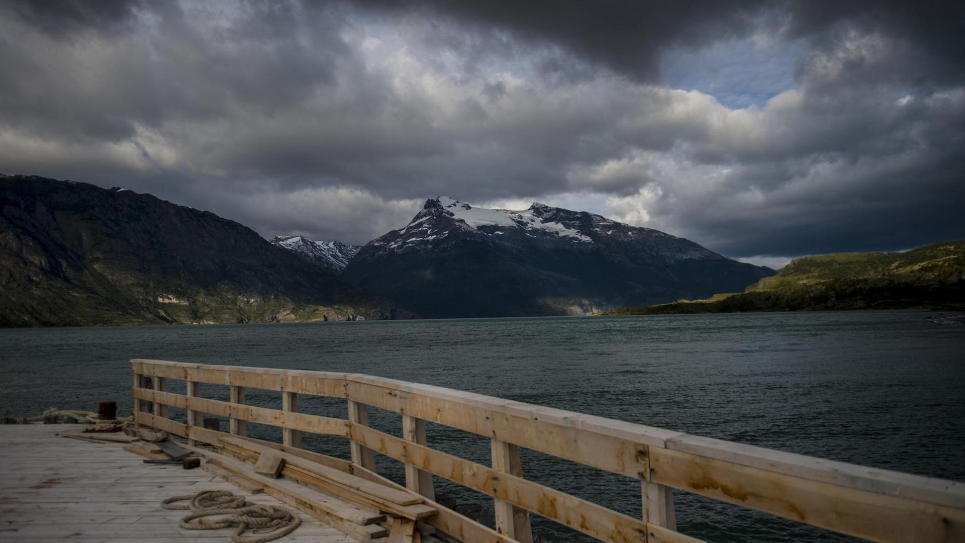 Blick von einem Holzsteg über einen Fjord, dahinter schneebedeckte Berge unter dunklen Wolken.