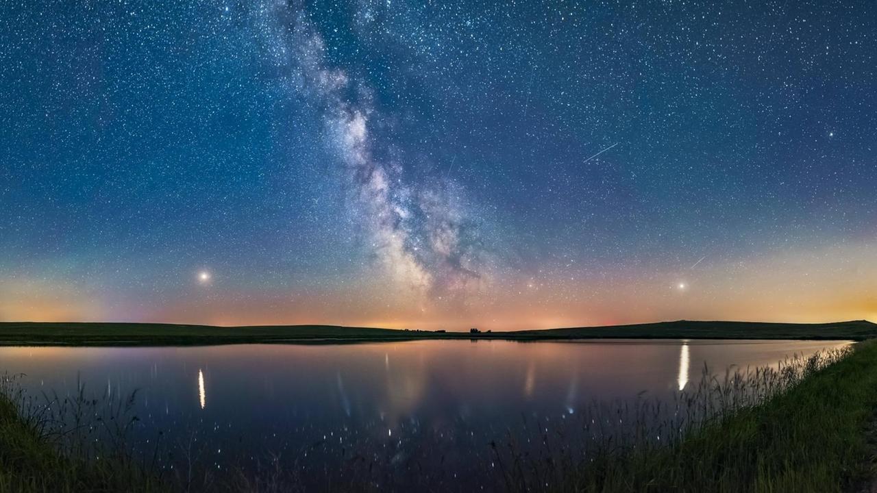 Panoramaaufnahme des nächtlichen Sternenhimmels mit Milchstraße über einem See.