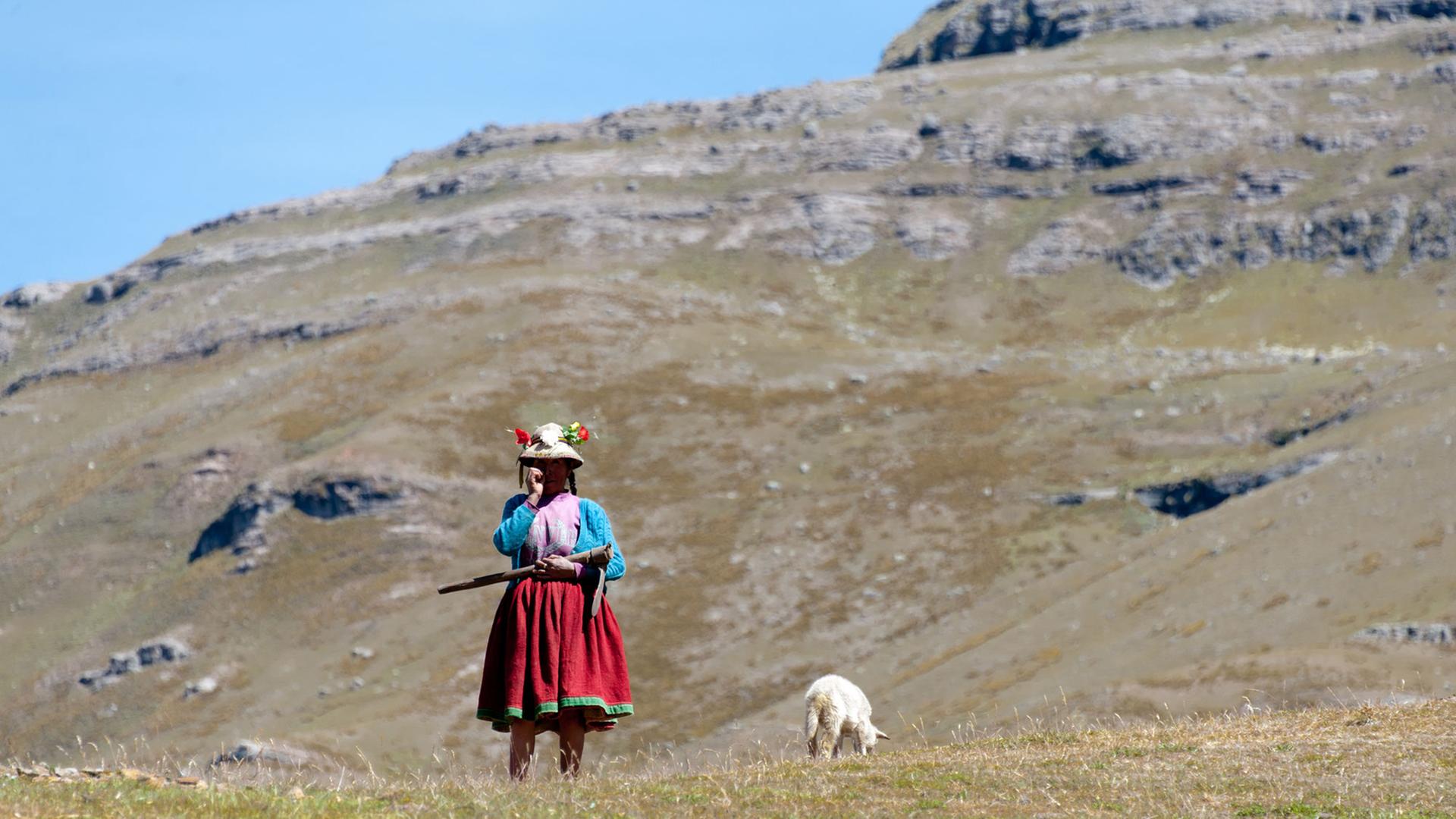 Eine Frau in traditioneller Kleidung steht vor einem Berpanorama in den peruanischen Anden.