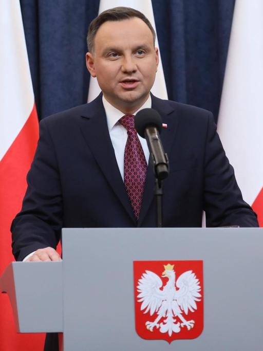 Duda steht an einem Rednerpult mit dem polnischen Adler und spricht. Hinter ihm vier polnische Flaggen vor einem blauen Vorhang.