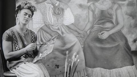 Eine Frau mit Farbkasten sitzt vor einem Gemälde. Es ist die Malerin Frida Kahlo.