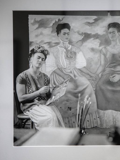 Eine Frau mit Farbkasten sitzt vor einem Gemälde. Es ist die Malerin Frida Kahlo.