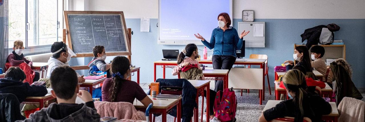 Schülerinnen und Schüler sitzen mit Masken und Abständen in einem Klassenraum. Vorne steht eine Lehrerin, ebenfalls mit Maske.
