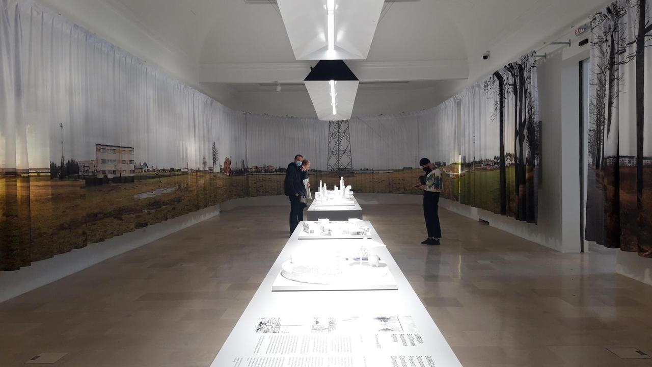 Blick in den polnischen Pavillon bei der 17. Architekturbiennale in Venedig. Entland den Wänder sind mit Landschaftsfotografien bedruckte Vorhänge aufgehängt und in der Mitte befindet sich ein langer Tische mit weißen Modellen und Informationstafeln.