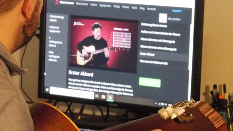 Overshoulder-Einstellung von Fardin Pfannkuche, der seine Gitarre hält und auf den Online-Videokurs auf seinem Laptop blickt.