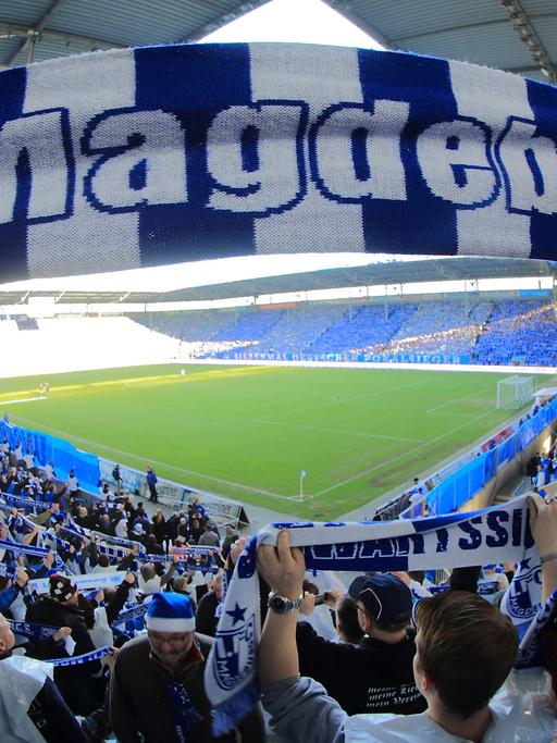 Ein Fan hält einen blau-weißen Schal mit der Aufschrift 1. FC Magdeburg in die Höhe, im Hintergrund sind die Ränge und der Rasen in einem Fußballstadion zu sehen.