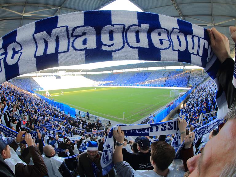 Ein Fan hält einen blau-weißen Schal mit der Aufschrift 1. FC Magdeburg in die Höhe, im Hintergrund sind die Ränge und der Rasen in einem Fußballstadion zu sehen.