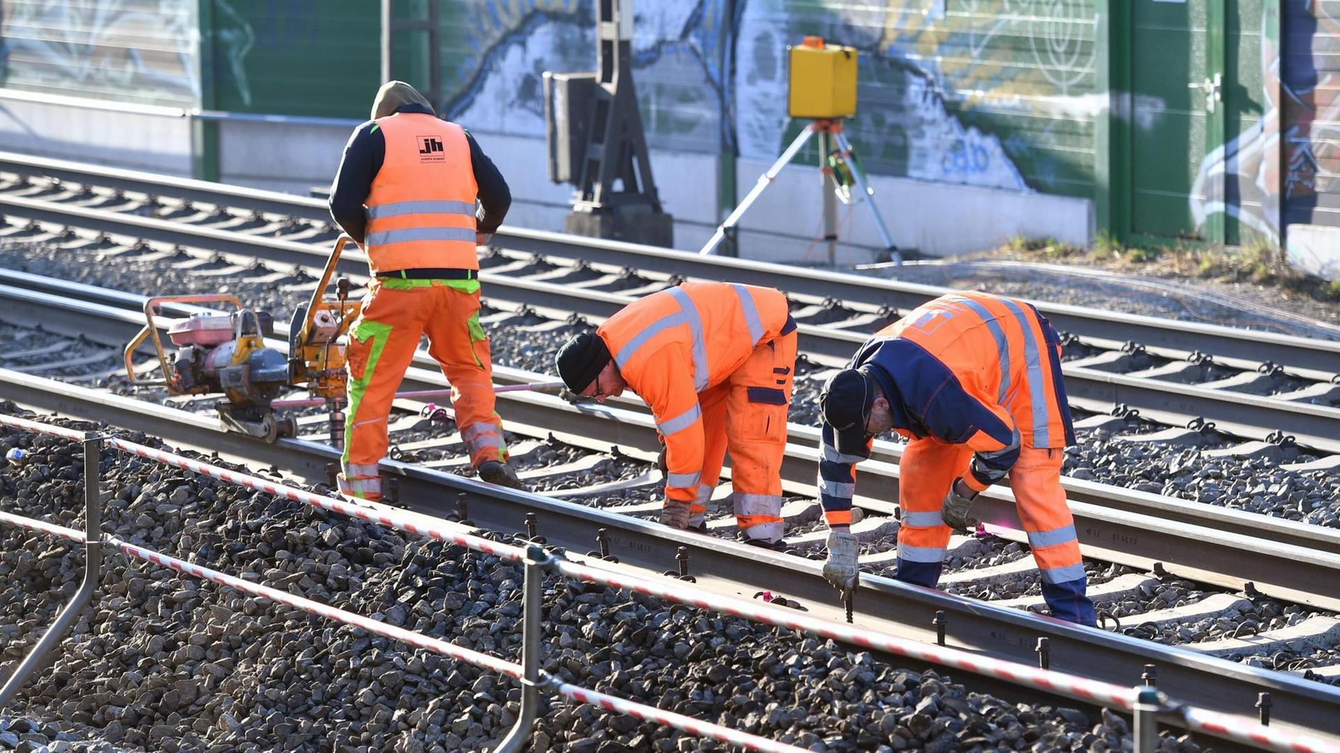 Drei Gleisarbeiter in roten Warnwesten und -hosen arbeiten an einer mehrgleisigen Strecke. Im Hintergrund eine Gebäudewand.