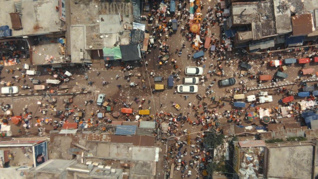 Ein Blick aus dem Luft nach unten zeigt eine wuselige Straßenszene in Indien. Mitten auf einer Kreuzung stehen drei weiße Autos. Fußgänger und andere motorisierte Fahrzeuge sind ebenso zu sehen wie die Dächer der anliegenden Häuser.