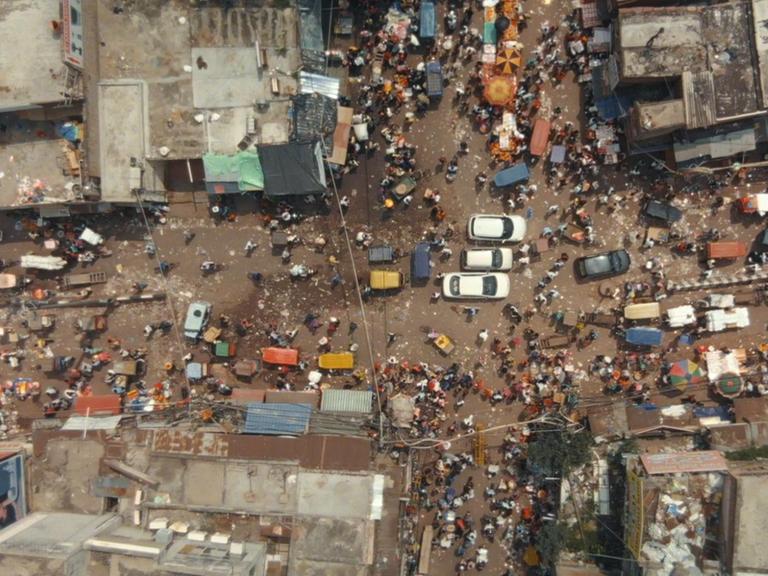 Ein Blick aus dem Luft nach unten zeigt eine wuselige Straßenszene in Indien. Mitten auf einer Kreuzung stehen drei weiße Autos. Fußgänger und andere motorisierte Fahrzeuge sind ebenso zu sehen wie die Dächer der anliegenden Häuser.