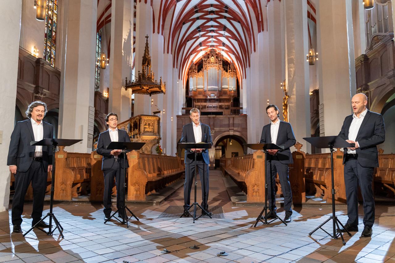 Fünf Sänger stehen hinter Notenständern inmitten der Kirche mit ihren rot gerandeten Gewölbebögen.