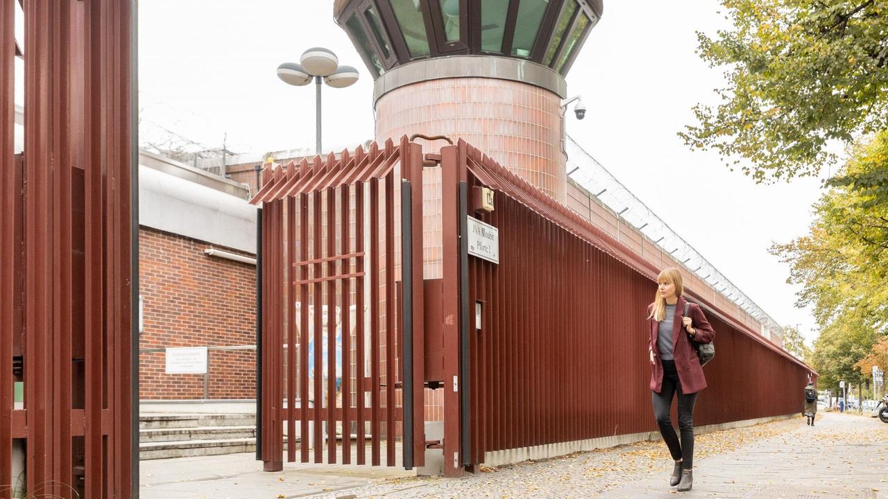 Eine junge Frau mit langen blonden Haaren und weinrotem Blazer geht an einer Gefängnismauer entlang auf ein großes offenes Tor zu.