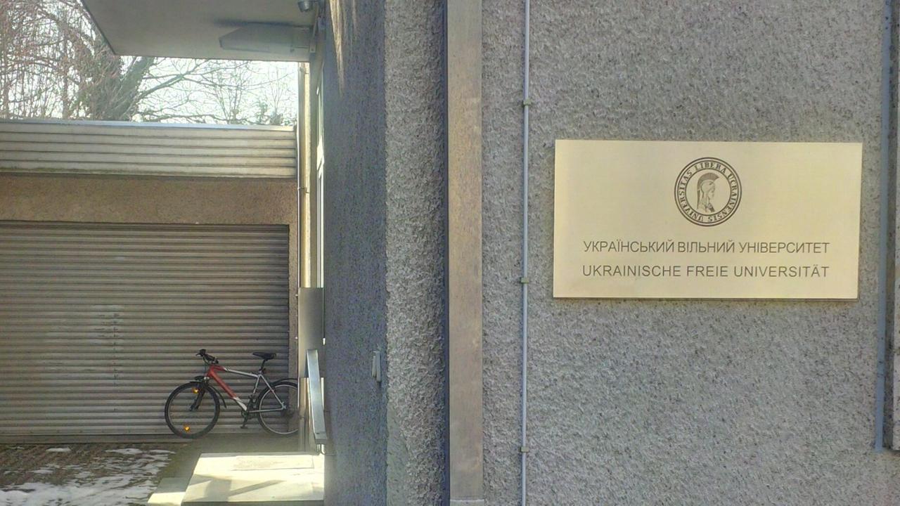Goldenes Schild mit Aufschrift "Ukrainische Freie Universität München", auch in Kyrillisch an einer grauen Hauswand, im Hintergrund steht ein Fahrrad an einer Wand.