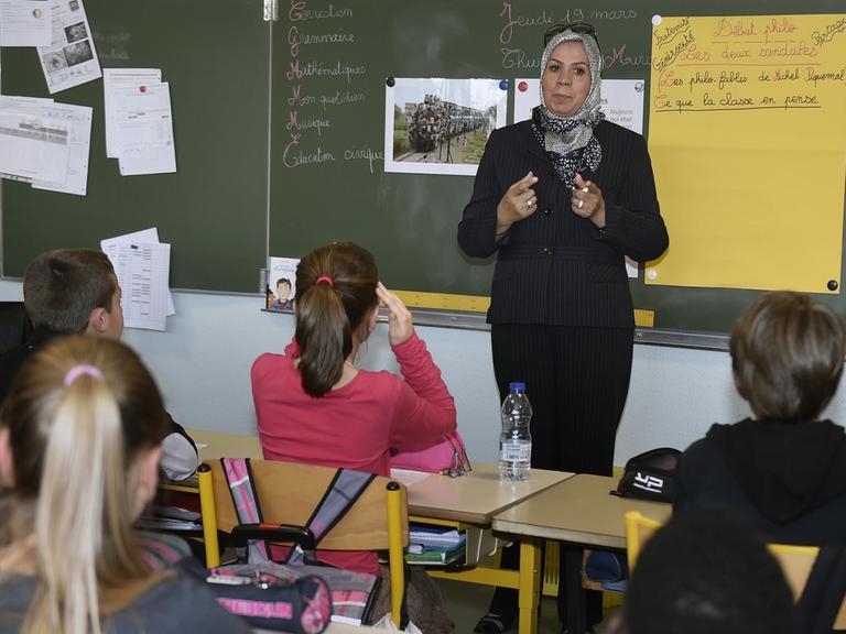 Eine Frau mit Kopftuch steht vor einer Schulklasse und spricht.