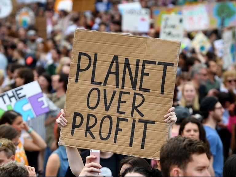 Bei den Protesten des Klimastreiks ist ein Schild zu sehen auf dem "Planet over Profits" zu lesen ist. Dahinter ist eine große Menschenmenge zu erkennen.