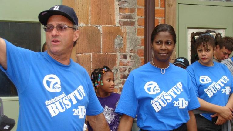 John George steht gemeinsam mit zwei Frauen - alle in knallblauen T-Shirts auf denen "Bustup" steht - bei einer öffentlichen Aktion in einer Reihe.