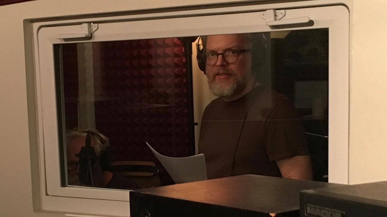 Der Podcast-Produzent Jonathan Mitchell steht mit Kopfhörern und Manuskript in einem Radio-Studio.