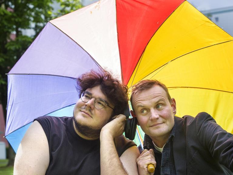 Devid Striesow und Axel Ranisch lehnen sich unter einem bunten Regenschirm aneinander und lauschen den Tönen aus einem Kopfhörer.