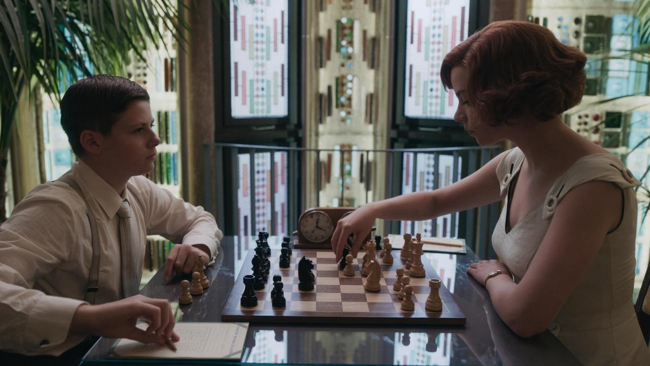 Anya Taylor-Joy, in der Rolle der  Elizabeth "Beth" Harmon,  sitzte am Tisch und spielt gegen einen jungen Mann Schach.