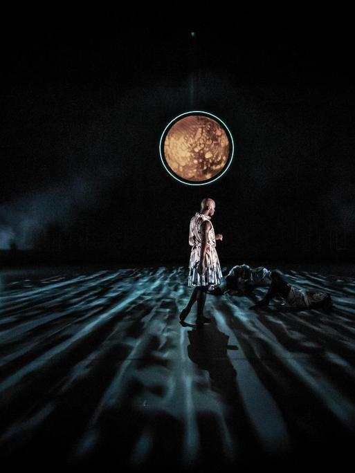 Szene aus dem Requiem: Ein Mann in einem Kleid steht auf einer mondbeschienenen Bühne. Am Boden liegen Tänzer.