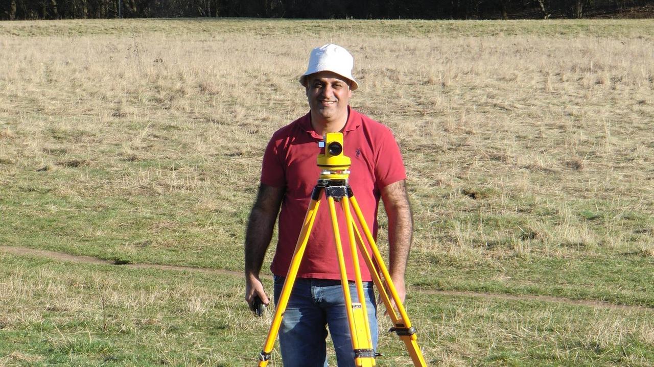 Ismael Ibraheem bei der Feldarbeit. Er steht auf einer Wiese vor sich seine Messapparatur und lacht in die Kamera. Die Sonne scheint, er trägt einen kleinen Sonnenhut.