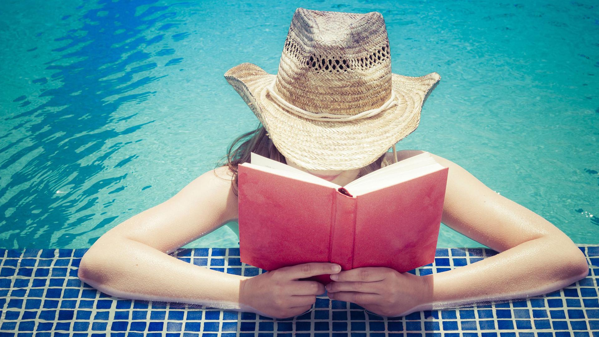 Eine Frau mit Strohhut liest am Beckenrand eines Schwimmbeckens ein Buch.