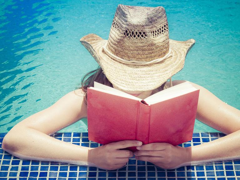 Eine Frau mit Strohhut liest am Beckenrand eines Schwimmbeckens ein Buch.