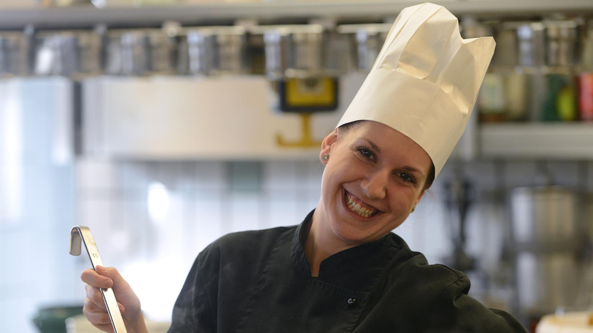 Eine Frau mit Kochmütze auf dem Kopf steht in einer Restaurantküche und rührt mit einem Löffel in einem Topf.