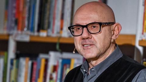 Andreas Pflüger steht vor einem Bücherregal in der Stadt- und Regionalbibliothek in Erfurt. Er hat eine Glatze, trägt eine Brille mit kräftigem, schwarzem Rand, hat ein graues Hemd und eine schwarze Weste an.