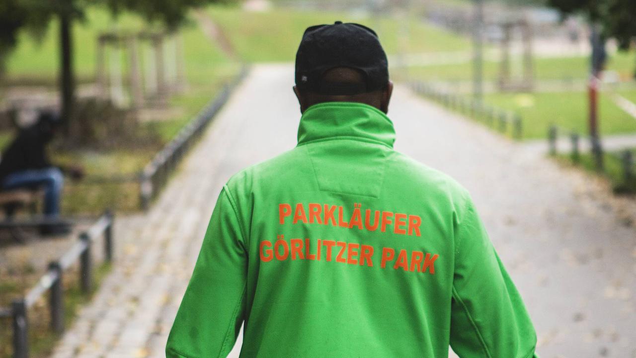 Mann sieht einen Mann von hinten in gründer Jacke, auf dem "Parkläufer" steht. Parkläufer arbeiten im Berliner Görlitzer Park als eine Art Sozialvermittler zwischen Drogendealern und Polizei.