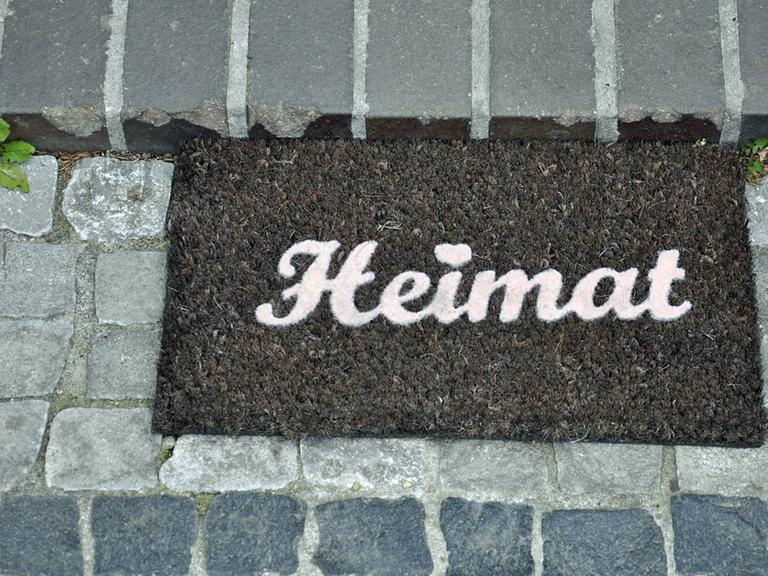 Ein Fußabtreter mit dem Schriftzug "Heimat"liegt auf einem gepflasterten Weg.