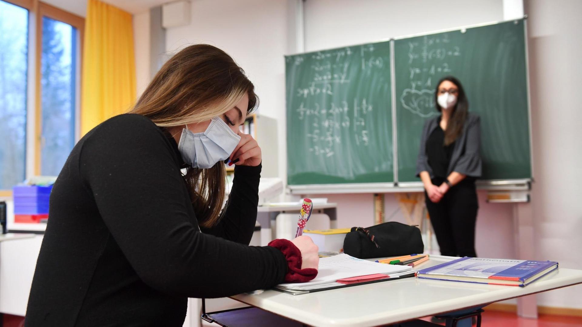 Schulunterricht in Zeiten der Coronavirus Pandemie: Lehrerin steht an einer Tafel, Schülerin arbeitet konzentriert.