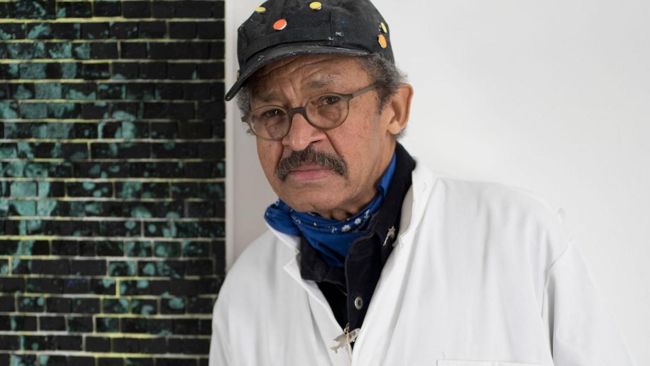 Der Künstler Jack Whitten mit Brille und Kappe lehnt gegen eine Wand.