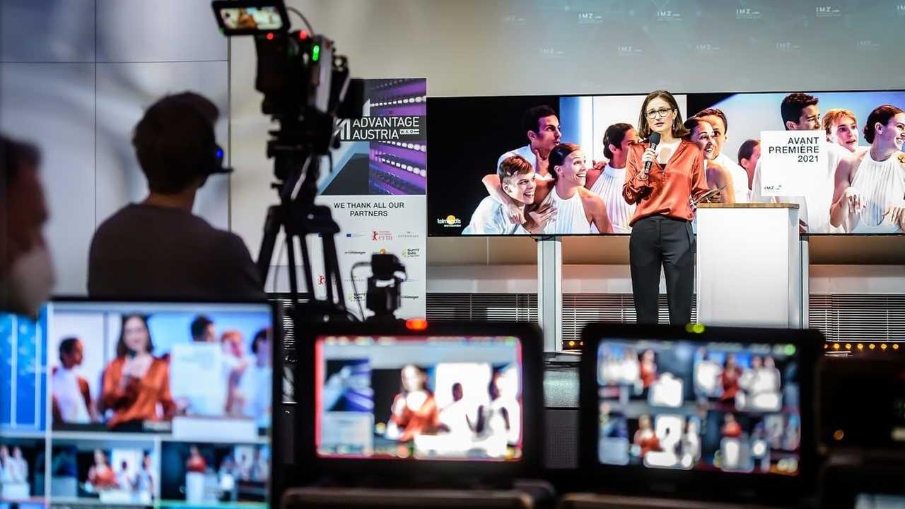Eine Frau steht vor Kameras in einem Studio und moderiert. Verschwommen im Vordergrund sind die Bildschirme der Bildregie zu sehen.