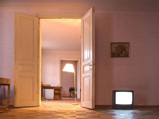 Eine halb leergeräumte Altbauwohnung, Möbel stehen in der Ecke und ein Fernseher leuchtet auf dem Fussboden.