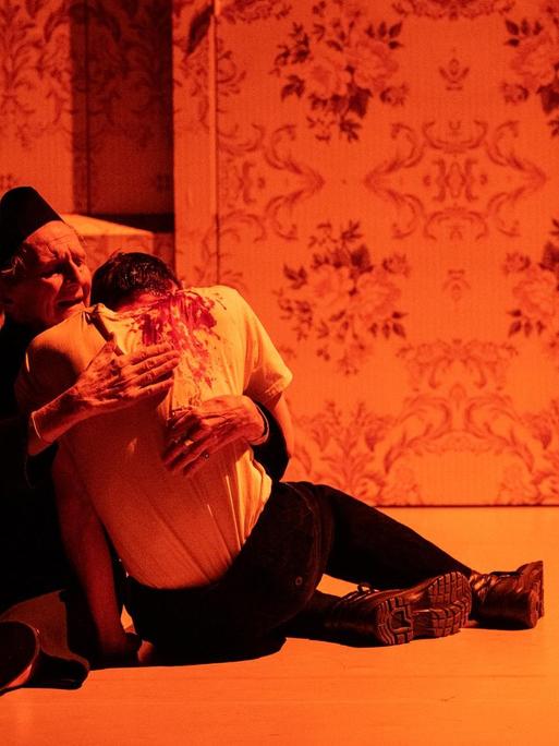 zwei Schauspieler umarmen sich sitzend, der eine scheint verletzt, er hat Blut an seinem Hemd. Die Szene ist in rotes Licht getaucht