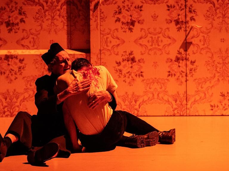 zwei Schauspieler umarmen sich sitzend, der eine scheint verletzt, er hat Blut an seinem Hemd. Die Szene ist in rotes Licht getaucht