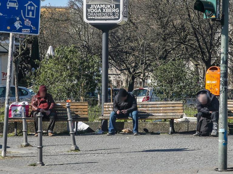 Drei Obdachlose sitzen in Berlin mit großem Abstand auf Parkbänken auf einem Platz in Kreuzberg.