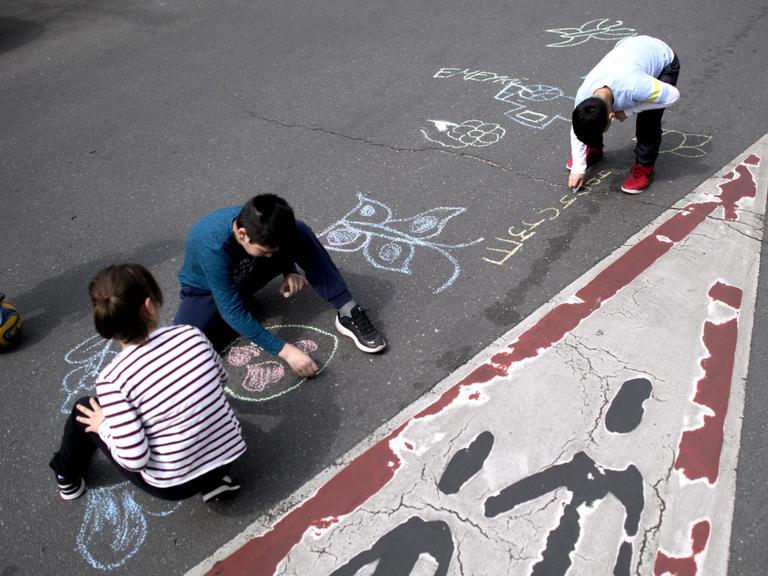 Kinder malen mit Kreide auf einer Straße, auf der sonst rund um die Uhr Autos fahren, ein Kind Kinder malen mit Kreide auf einer Straße, auf der sonst rund um die Uhr Autos fahren.hält einen Ball unter seinem Fuß.