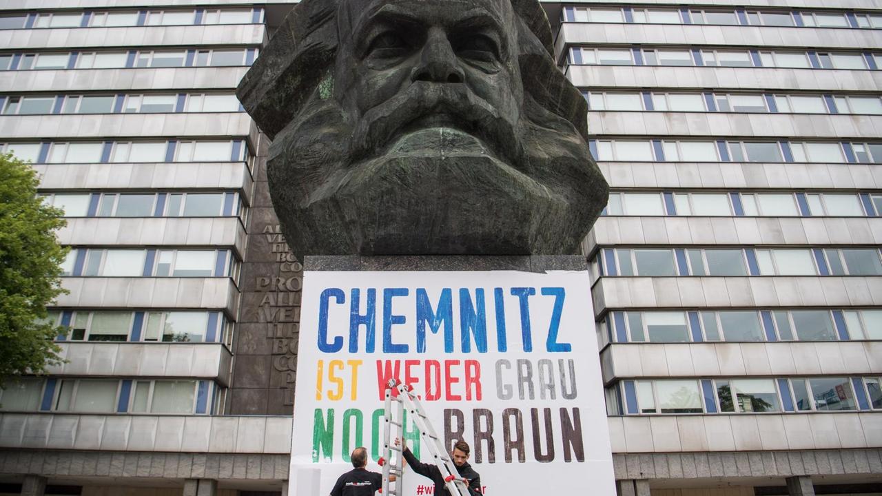 Zwei Männer befestigen am Karl-Marx-Denkmal ein Plakat mit der Aufschrift "Chemnitz ist weder grau noch braun". 