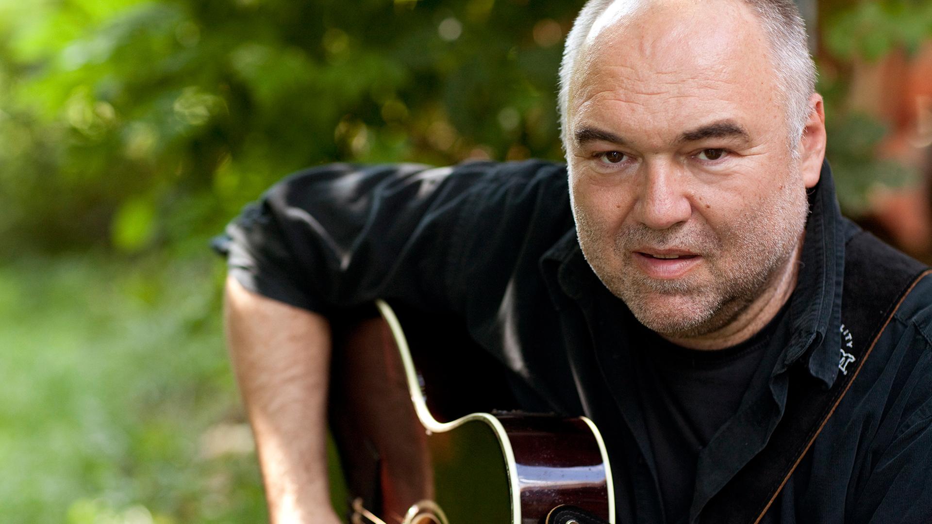 Zu sehen ist der oberfränkische Songpoet Wolfgang Buck sitzend und in die Kamera blickend mit einer Gitarre spielbereit unter dem rechten Arm.