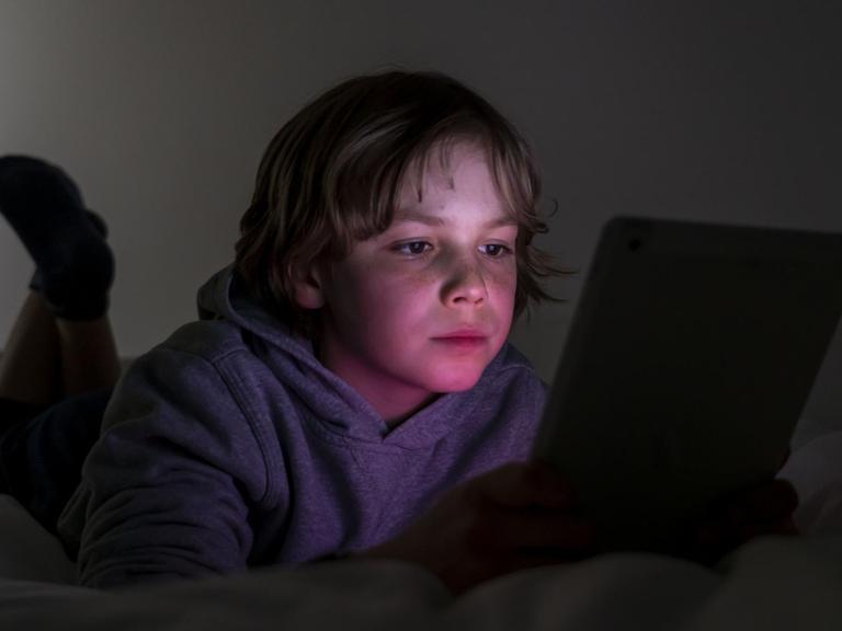 Ein Junge liegt auf einem Bett und spielt Computerspiele an einem Tablet.