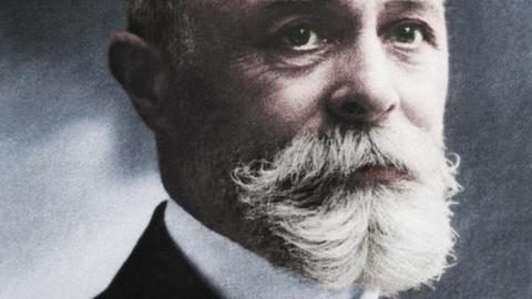 Die digitale Kolorierung eines Schwarzweiß-Fotos von 1903 zeigt den französischen Physiker Henri Becquerel mit weißen Vollbart, Glatze, und dunkelblauem Jackett über grüner Weste.